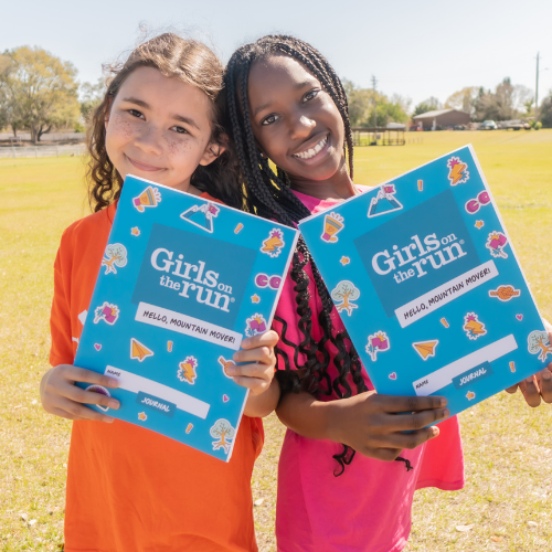 Two young girls reading a pink Girls on the Run journal outside.